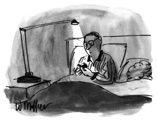 Man sitting in bed reading a very small book underneath a lamp on the bedside table next to him.