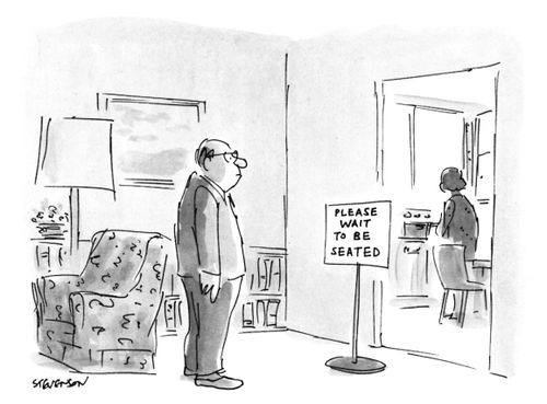 A man waits near a sign outside his kitchen that reads "Please wait to be seated" while his wife prepares dinner.