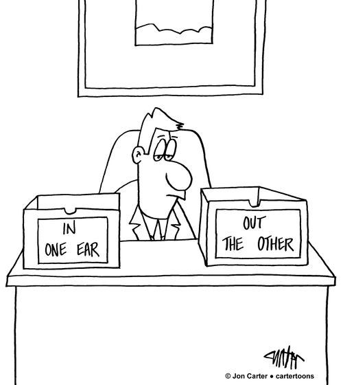 Man at desk has a desk tray labeled "In one ear,' and another labeled "Out the other."