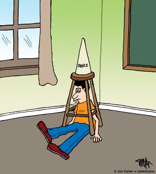 Student sits under a stool with a dunce cap on it in a classroom.