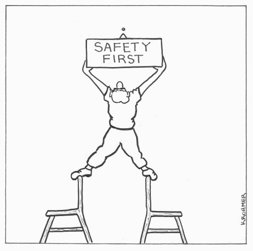 A man is balancing on two chairs to hang a 'safety first' sign.